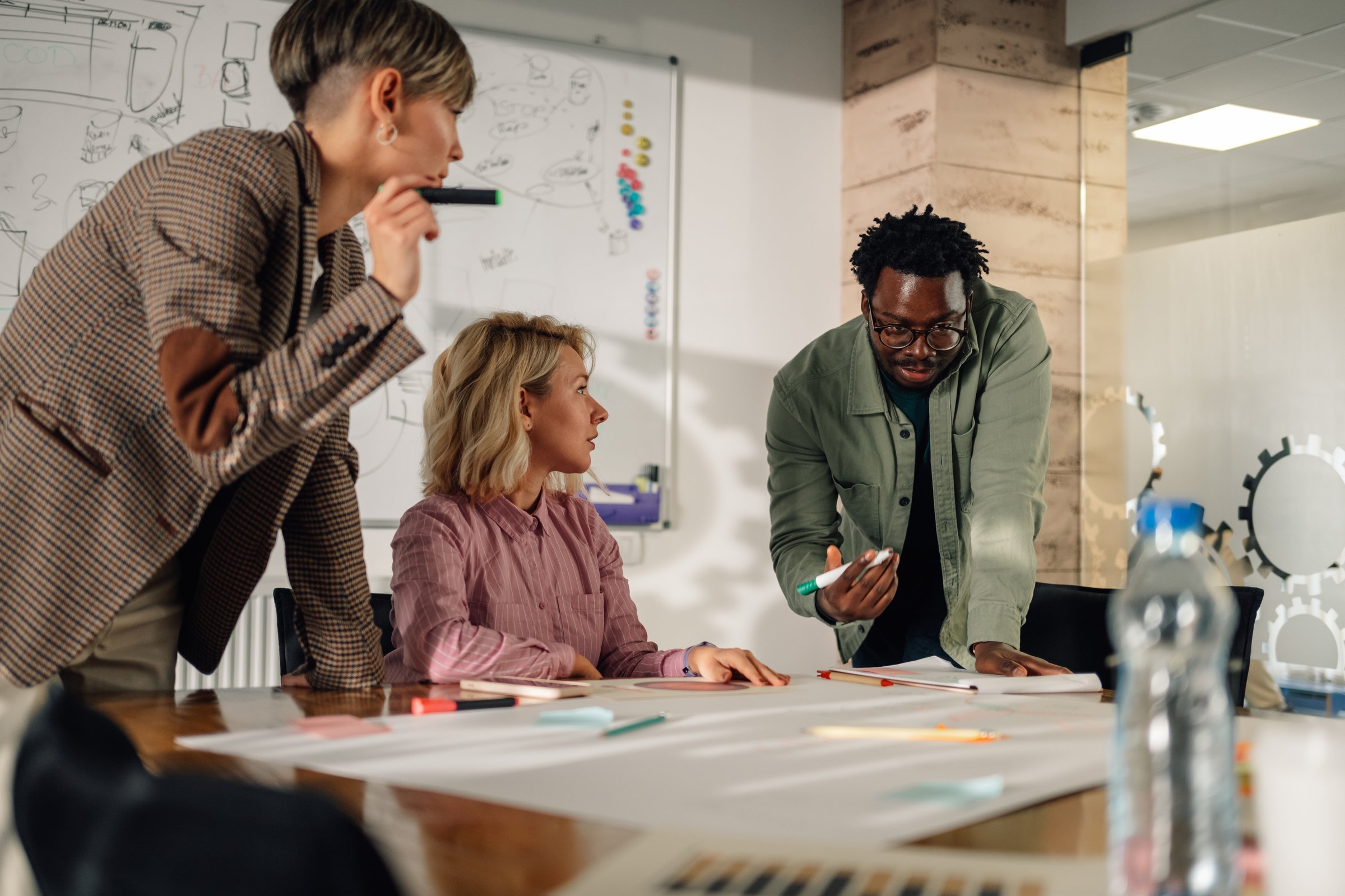 Three marketing specialists are working together on a project, analyzing diagrams and graphs spread out on a table during a productive meeting in a modern office